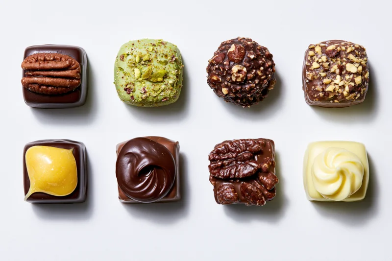 Twelve assorted artisan chocolate confections arranged in two rows on a white surface