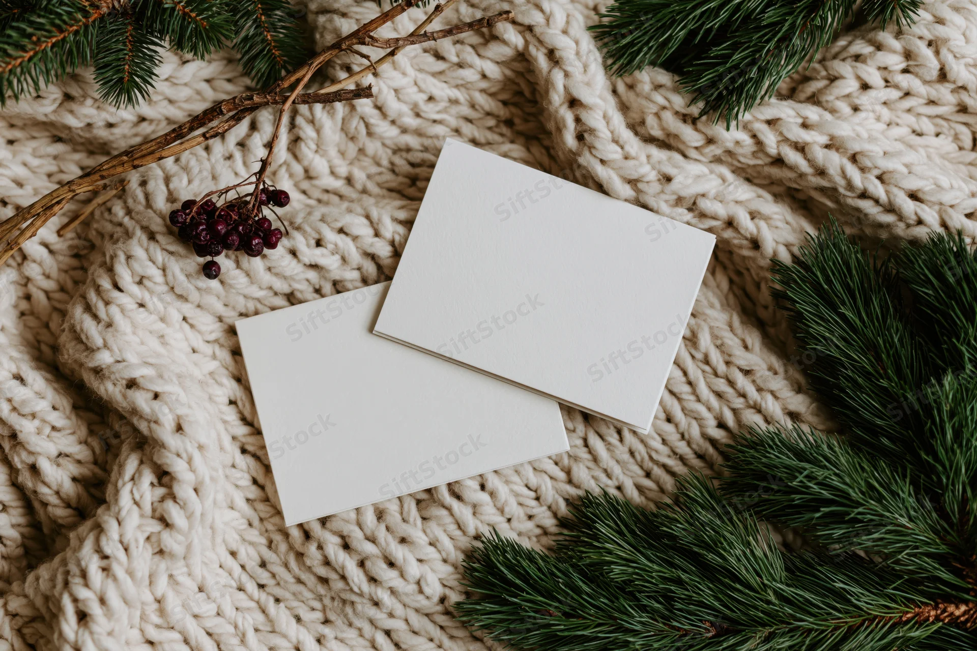 Two blank white greeting cards overlapping on a knitted wool sweater with pine branches and berries