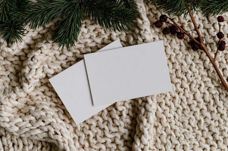 Two blank white greeting cards overlapping on a cream knit sweater with pine branches and dried berries