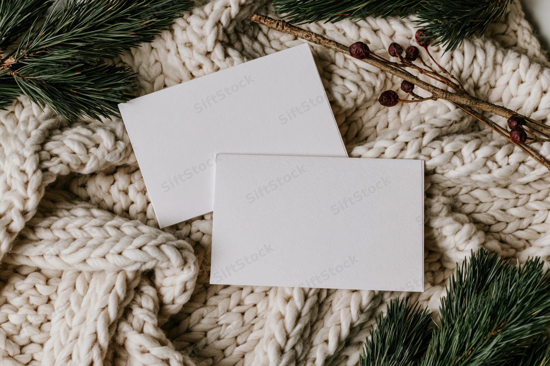 Two blank white greeting cards slightly overlapping on a chunky knit blanket with pine branches and berries