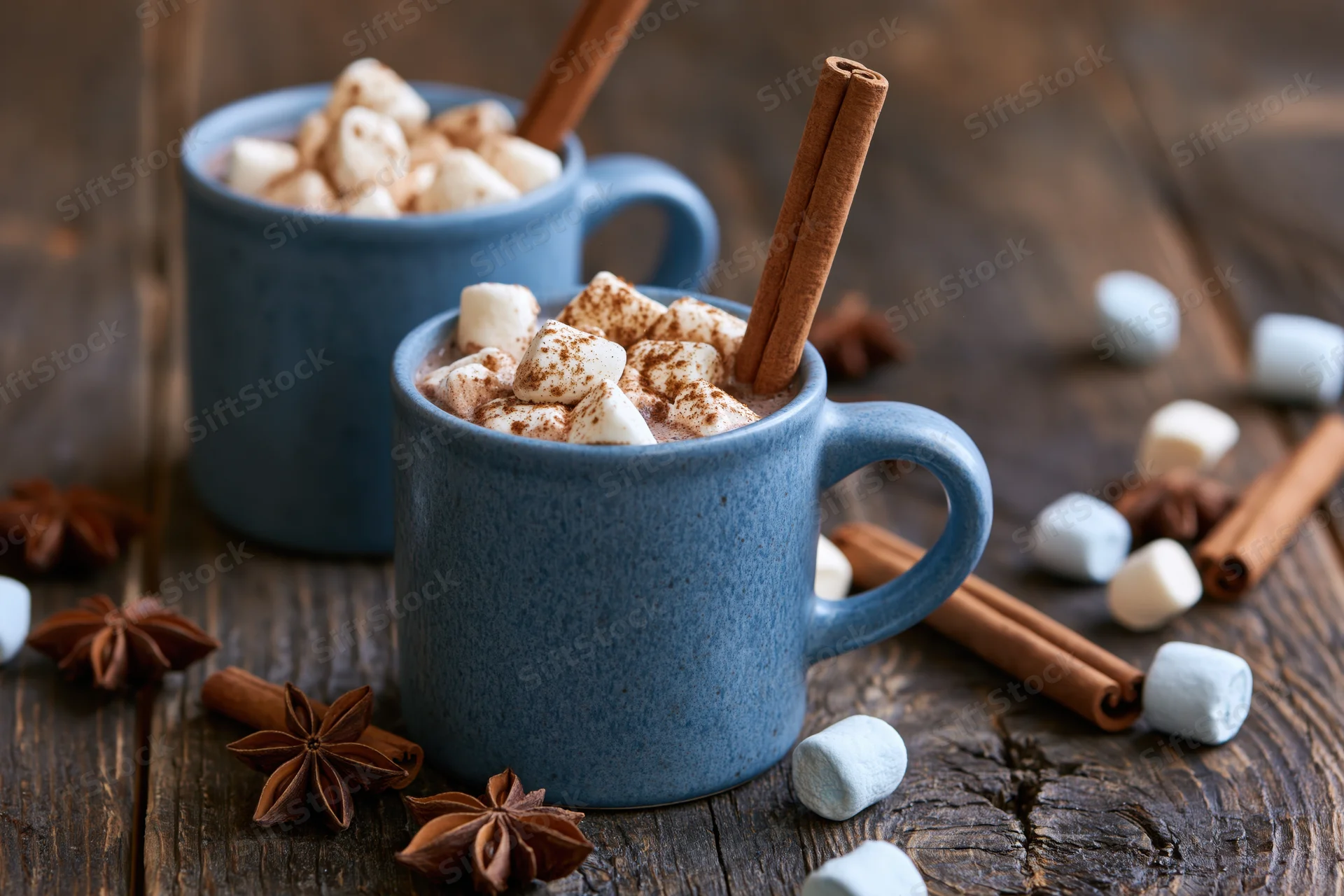 Two blue ceramic mugs filled with hot chocolate topped with marshmallows and cinnamon sticks on a wooden table