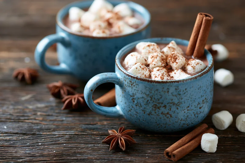 Two blue ceramic mugs filled with hot chocolate topped with marshmallows and cinnamon sticks on a wooden table