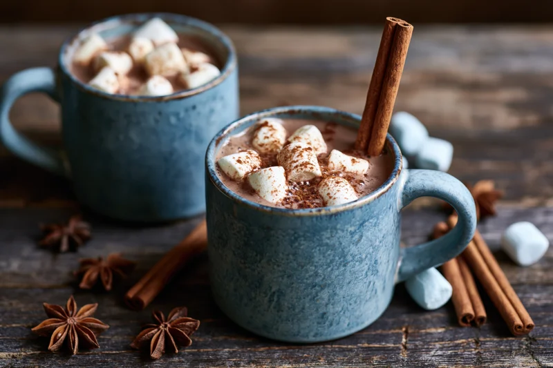 Two blue ceramic mugs filled with hot chocolate topped with marshmallows and cinnamon sticks on a wooden table