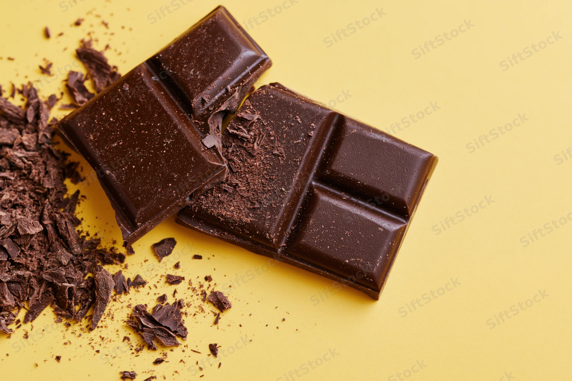 Broken dark chocolate bar pieces with scattered crumbs on yellow background
