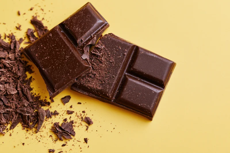 Broken dark chocolate bar pieces with scattered crumbs on yellow background