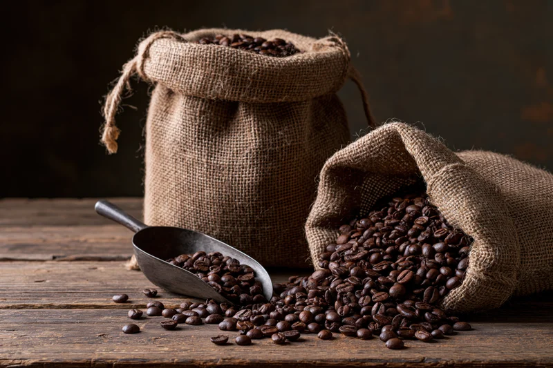 Roasted coffee beans spilling from burlap sacks onto a worn wooden table with a metal scoop nearby