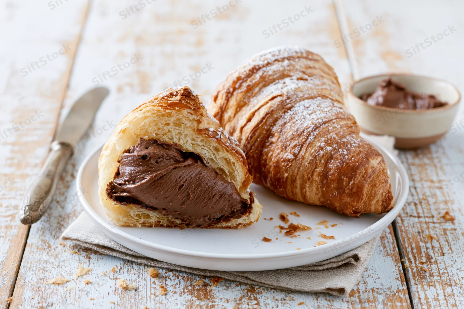 Two butter croissants on a white plate one sliced open with chocolate filling visible on a wooden surface