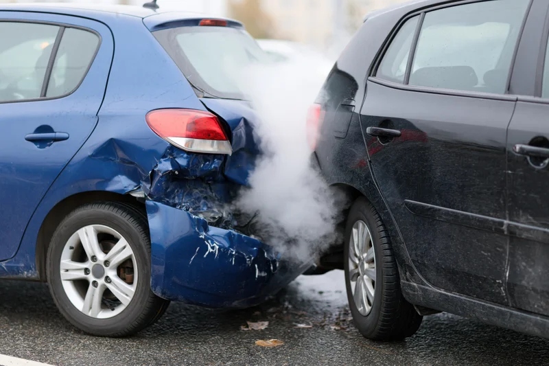 Two cars involved in a rear-end collision on a wet road with visible damage and smoke from the impact