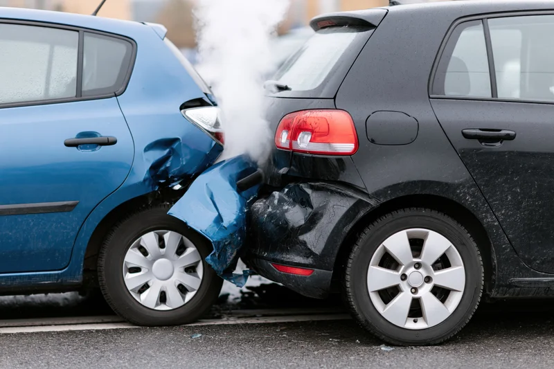 Two hatchback cars involved in a rear-end collision on a road with visible damage and smoke from the impact