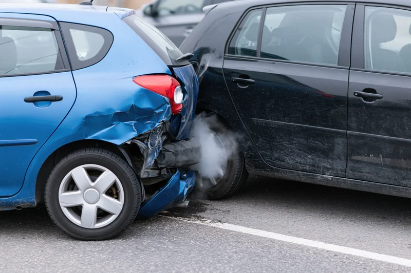 Two cars involved in a rear-end collision on a paved road with visible damage and smoke from the impact