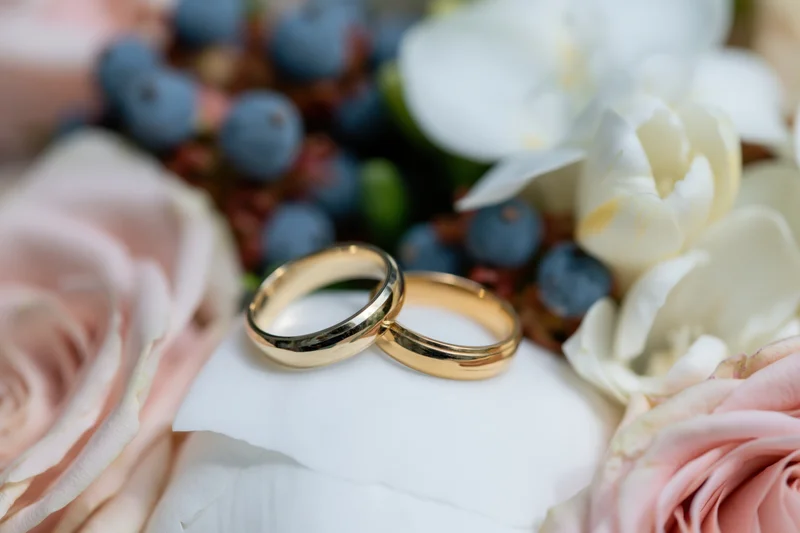 Two gold wedding bands resting side by side on a white peony surrounded by pink roses and blueberries