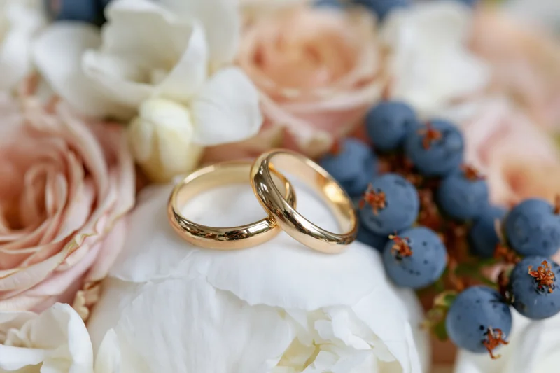 Two gold wedding bands resting side by side on a white peony surrounded by pink roses and blueberries
