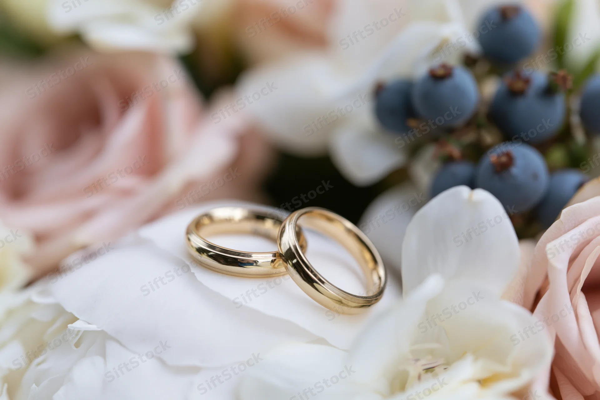 Two gold wedding bands resting side by side on a white peony petal within a floral bouquet