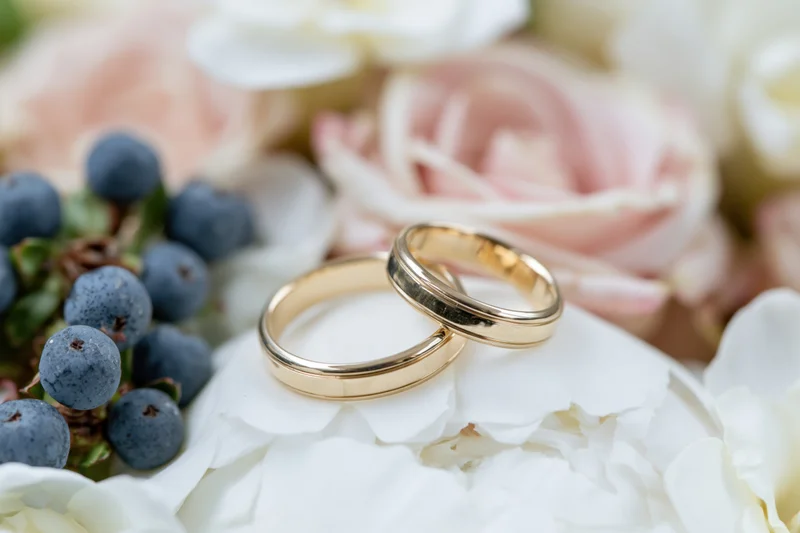 Two gold wedding bands resting side by side on a white peony flower among soft pink roses and blueberries