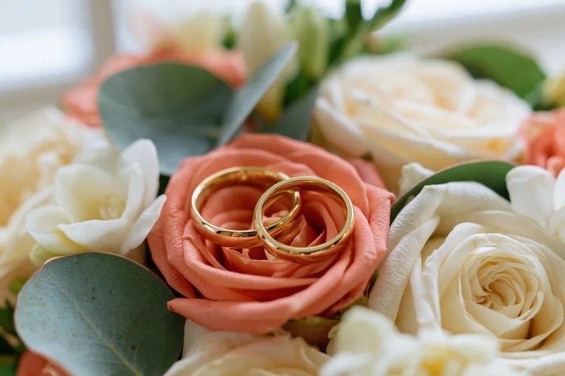 Two gold wedding rings resting on a bridal bouquet of coral and white roses with eucalyptus leaves