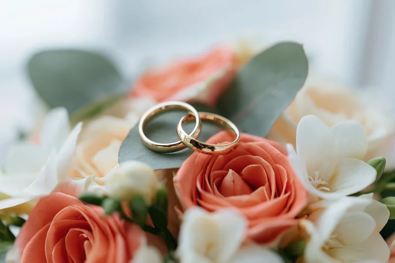 Two gold wedding rings resting on a bridal bouquet of coral and white roses with eucalyptus leaves