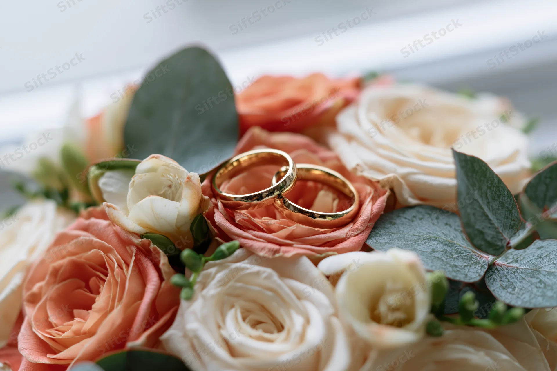 Two gold wedding rings resting on a bridal bouquet of coral and white roses with eucalyptus leaves