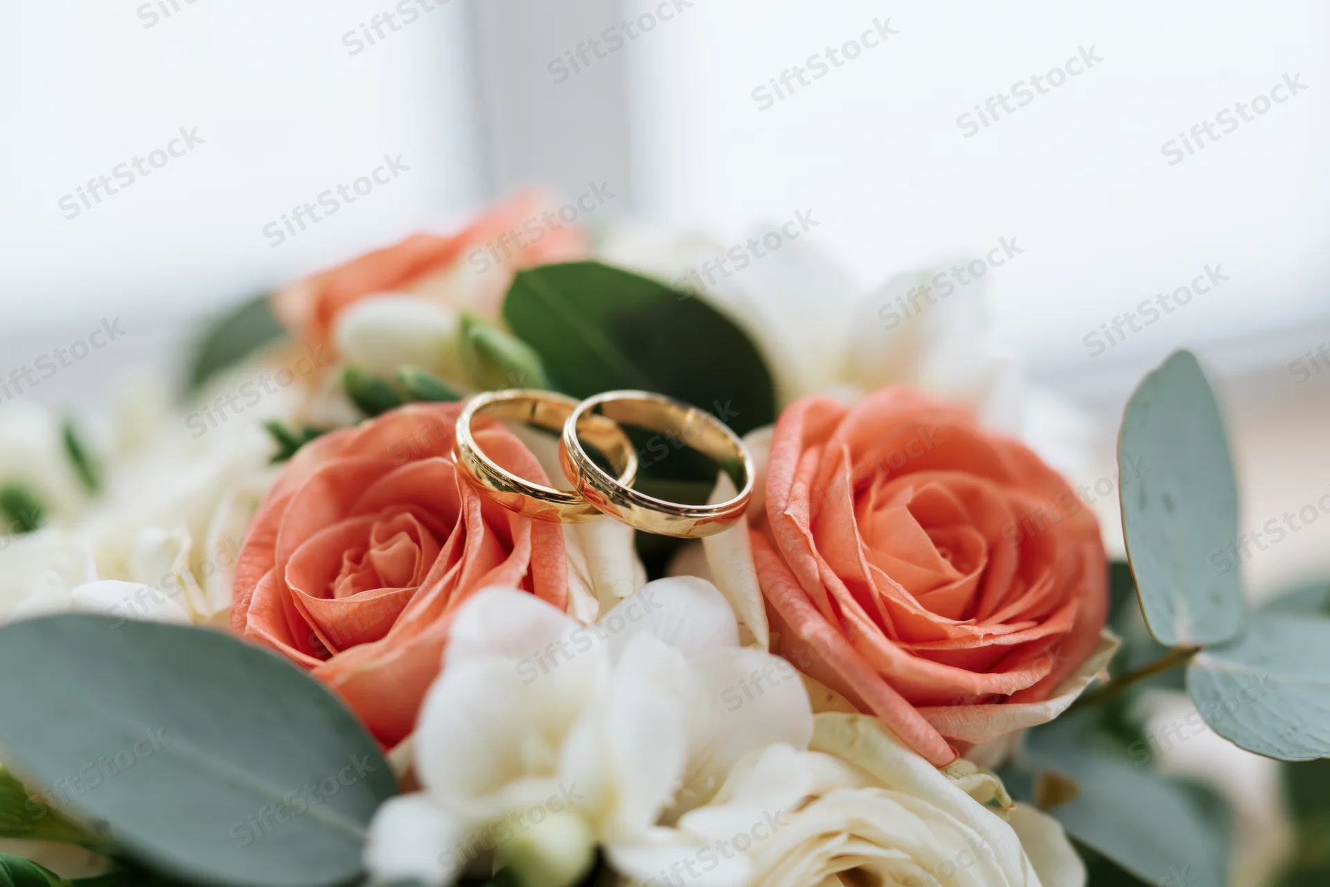 Two gold wedding rings resting on a bridal bouquet of coral and white roses with eucalyptus leaves