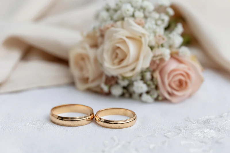 Two gold wedding rings resting side by side on white lace fabric with a floral bouquet in the background