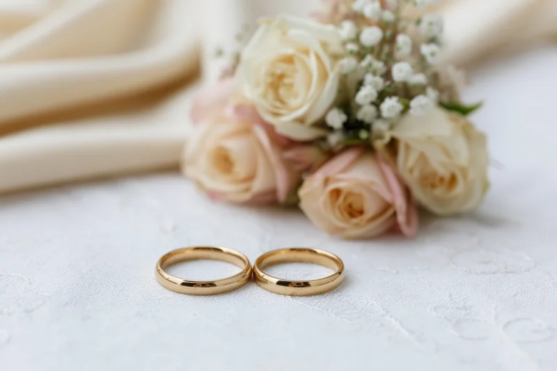 Two gold wedding rings resting side by side on white embroidered fabric with a floral bouquet in the background