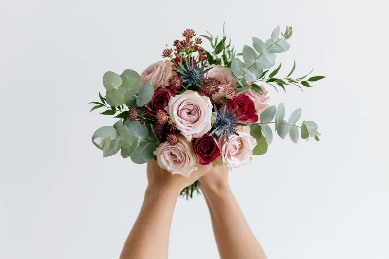 Hands holding a mixed floral bouquet of pink and red roses with eucalyptus greenery against a white background