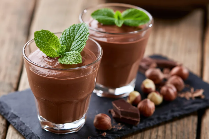 Two glasses of chocolate mousse garnished with mint leaves on a slate board with hazelnuts and chocolate pieces nearby