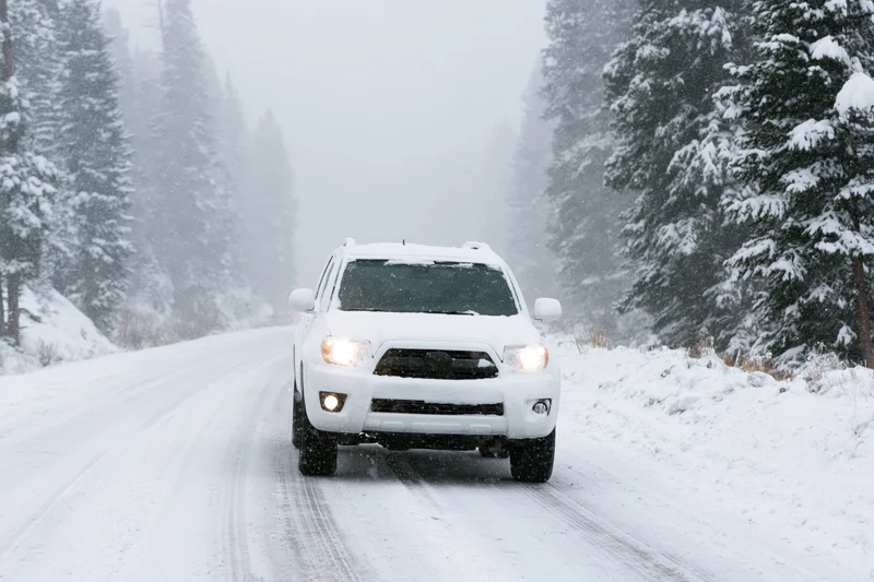 White suv driving on snow-covered mountain road surrounded by snow-laden evergreen trees during snowfall