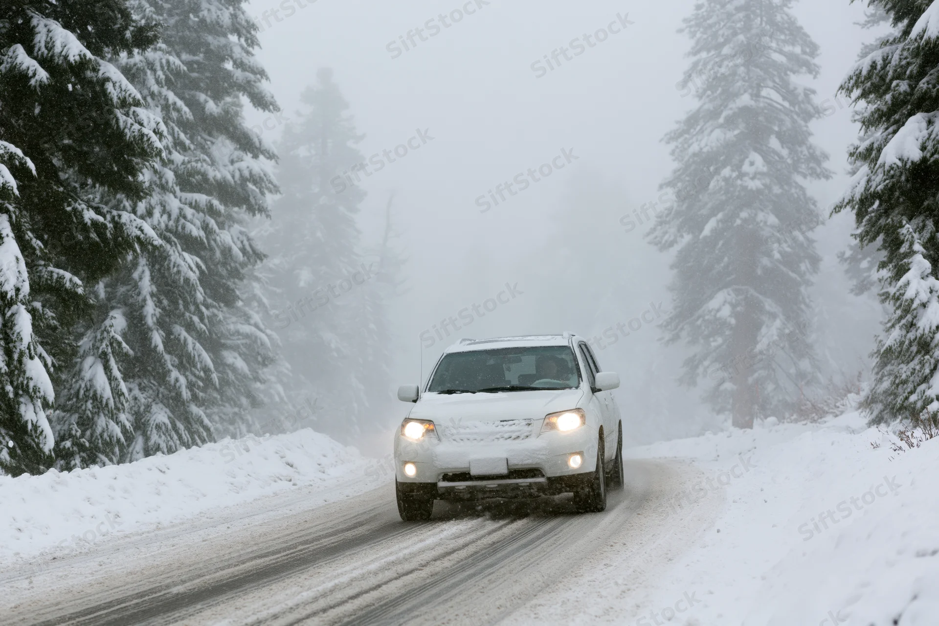 White suv driving on snow-covered mountain road through snowy forest with headlights on