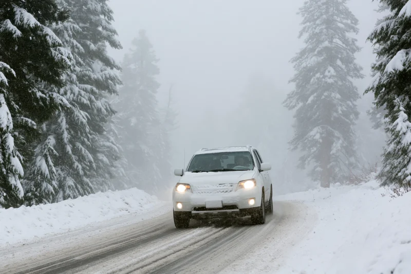 White suv driving on snow-covered mountain road through snowy forest with headlights on