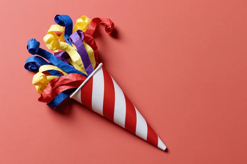 Red and white striped party popper cone lying on its side with colorful paper streamers spilling out on a coral background