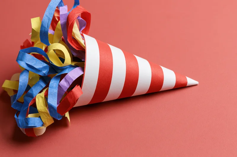 Red and white striped party popper cone lying on its side with colorful streamers spilling out on a solid red background