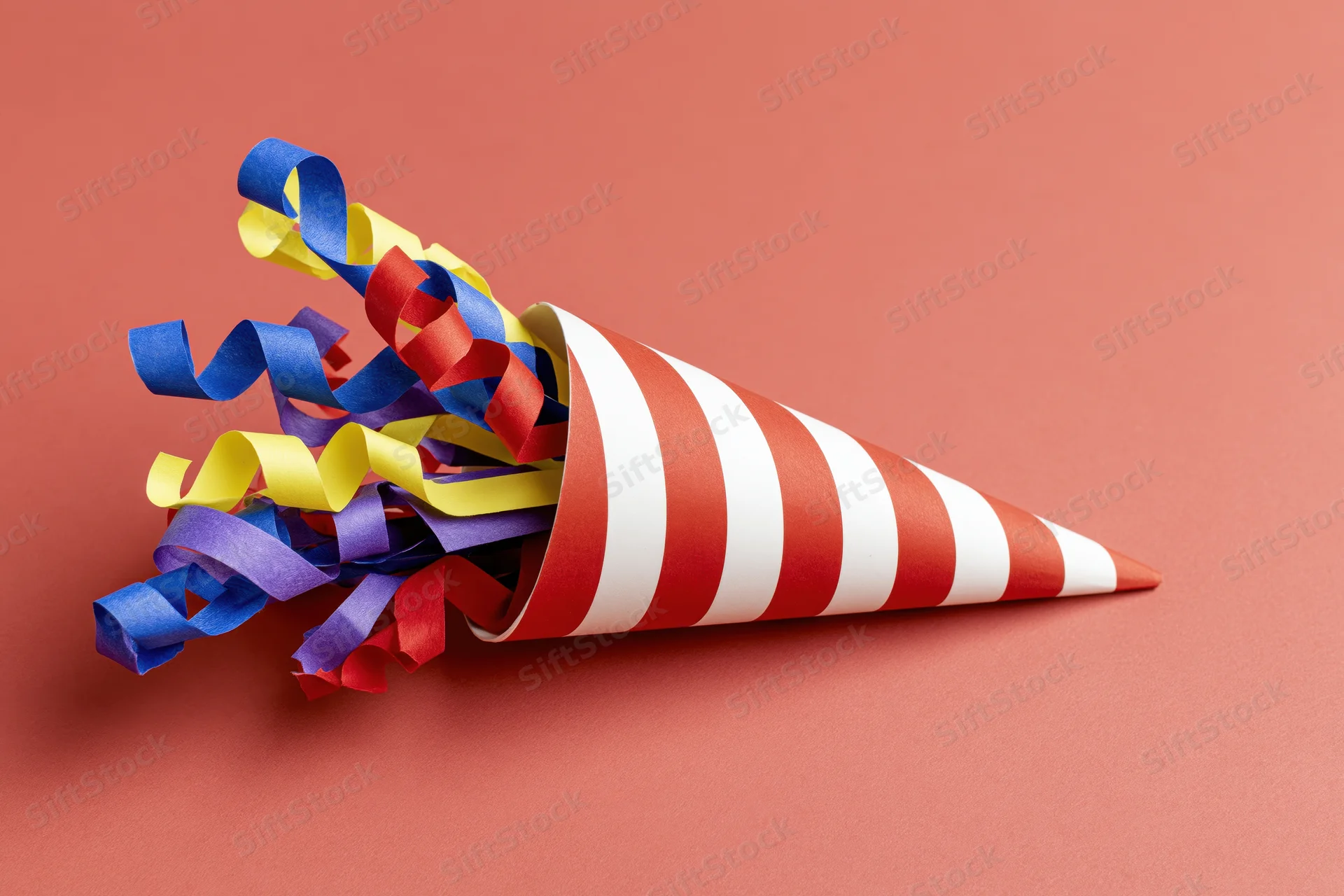Red and white striped party popper cone lying on its side with colorful ribbons spilling out on a solid coral background