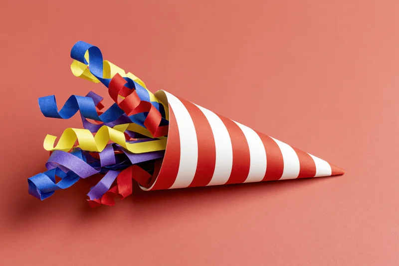 Red and white striped party popper cone lying on its side with colorful ribbons spilling out on a solid coral background