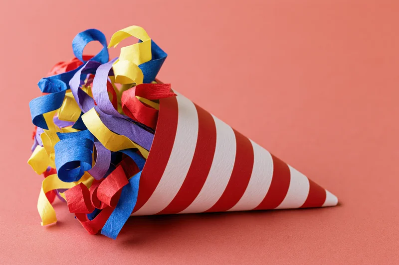 Red and white striped party popper cone lying on its side with colorful streamers spilling out on a solid peach background