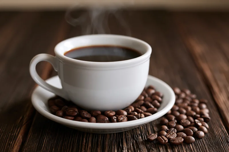 White ceramic coffee cup filled with steaming black coffee on saucer surrounded by roasted beans on dark wooden table