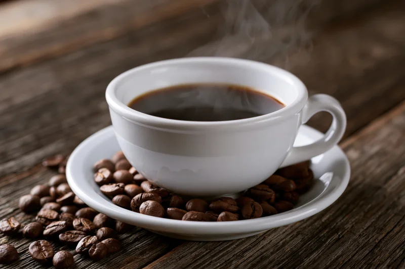 White ceramic coffee cup filled with steaming black coffee on saucer surrounded by roasted beans on wooden table