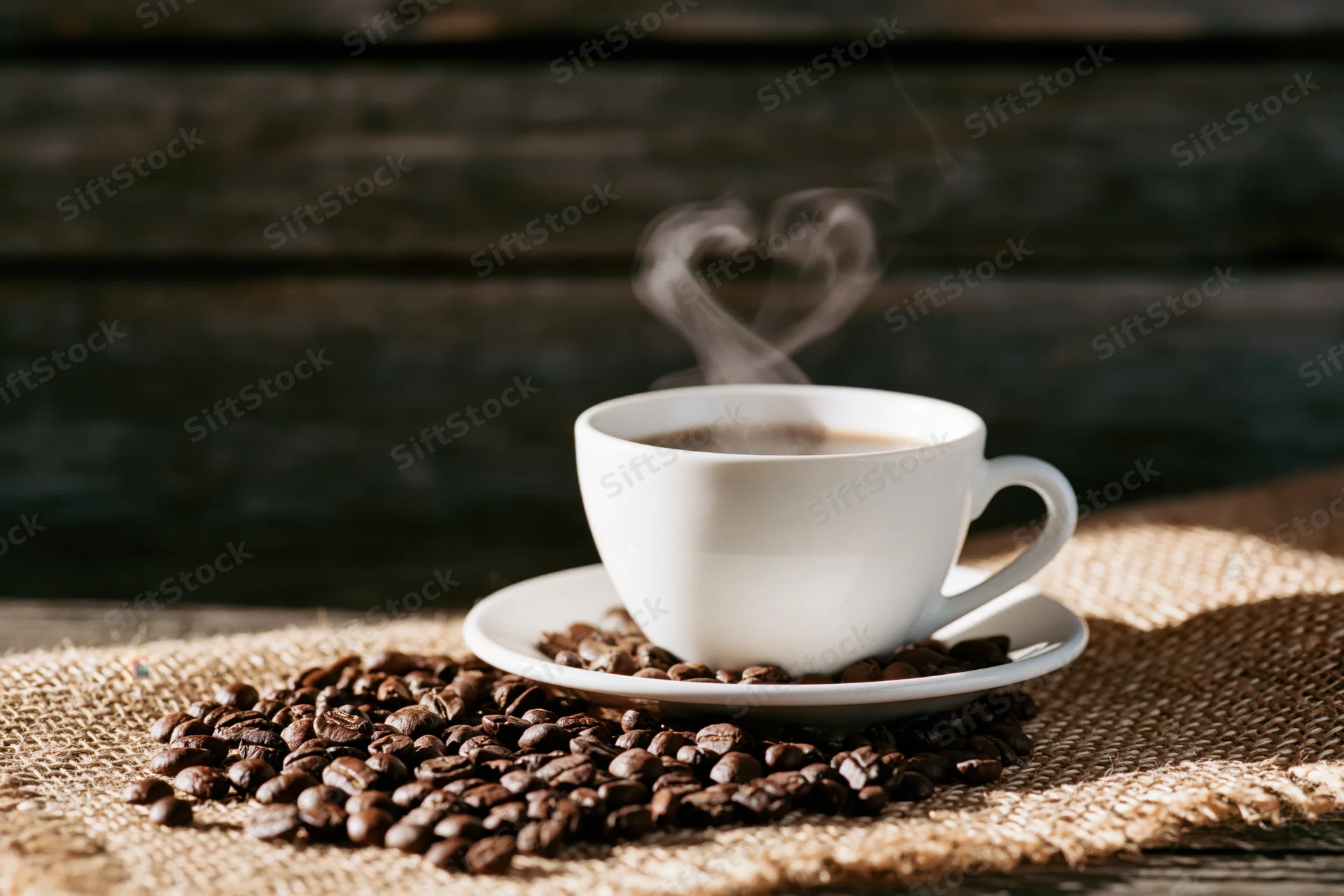 White ceramic coffee cup on saucer with heart-shaped steam rising over coffee beans on burlap cloth