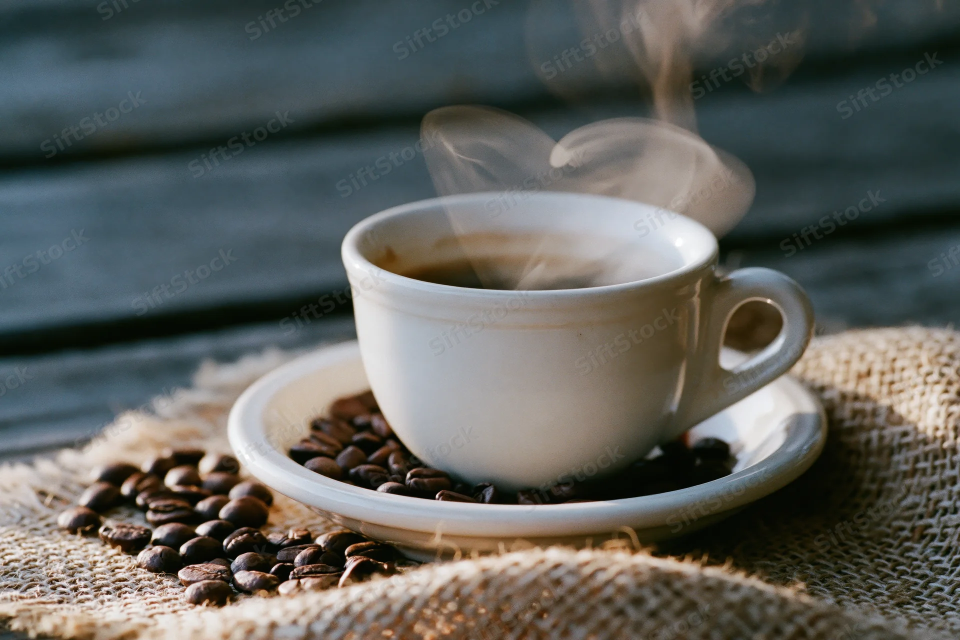 White ceramic coffee cup steaming on saucer with coffee beans scattered on burlap cloth over wooden surface