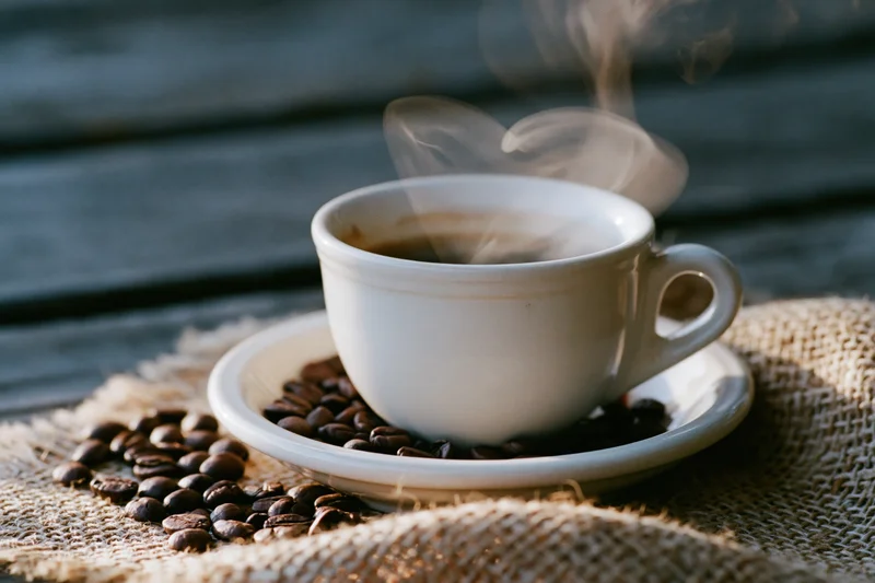White ceramic coffee cup steaming on saucer with coffee beans scattered on burlap cloth over wooden surface