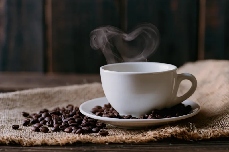 White ceramic coffee cup on saucer with heart-shaped steam rising, surrounded by coffee beans on burlap cloth over wooden table
