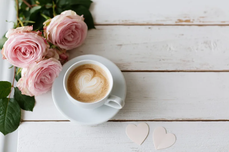 White ceramic cup of cappuccino with heart latte art on whitewashed wooden table beside pink roses and paper hearts