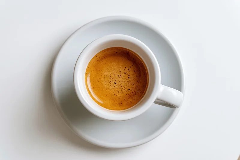White ceramic espresso cup filled with coffee and crema on matching saucer viewed from above on plain white surface