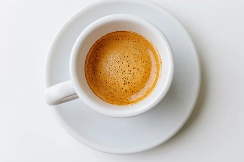 White ceramic espresso cup filled with coffee and crema on matching saucer against plain white background