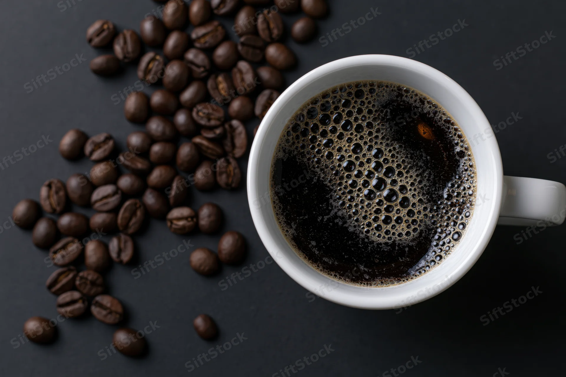 White ceramic mug filled with black coffee showing golden foam, scattered roasted beans on dark surface, top view