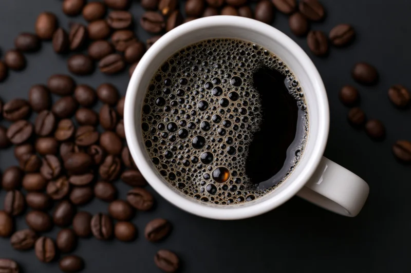 White ceramic mug filled with black coffee topped with golden foam surrounded by roasted coffee beans on dark surface