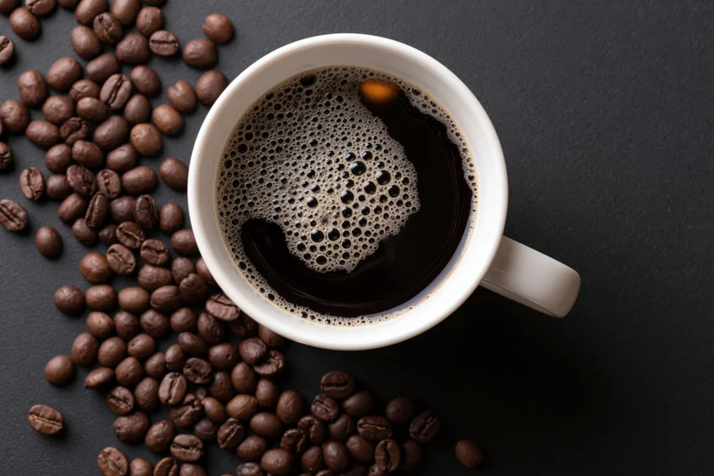 White ceramic mug filled with black coffee showing golden foam and crema, surrounded by roasted coffee beans on dark surface, top view
