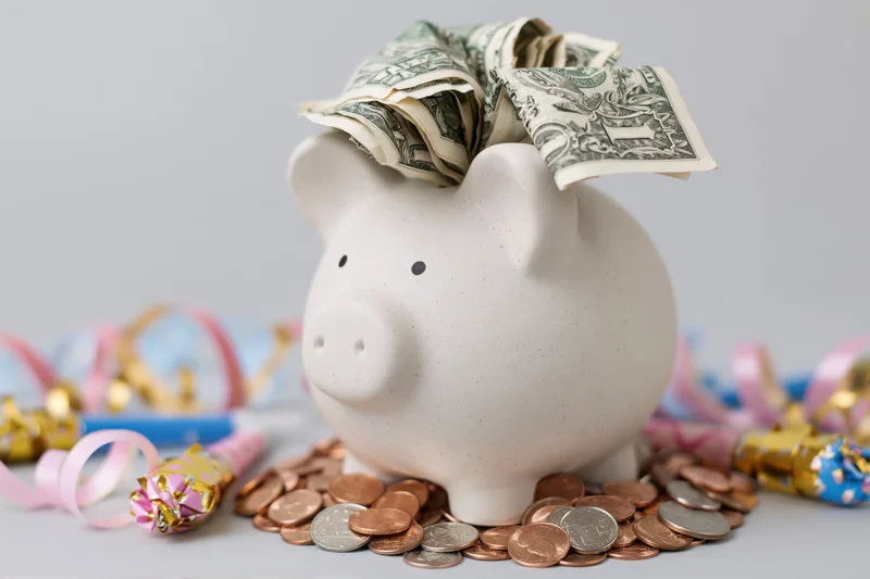 White ceramic piggy bank stuffed with folded us dollar bills surrounded by coins and festive decorations on a plain surface