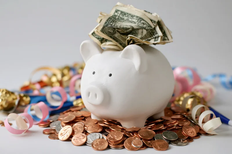 White ceramic piggy bank stuffed with folded us dollar bills surrounded by coins and party streamers