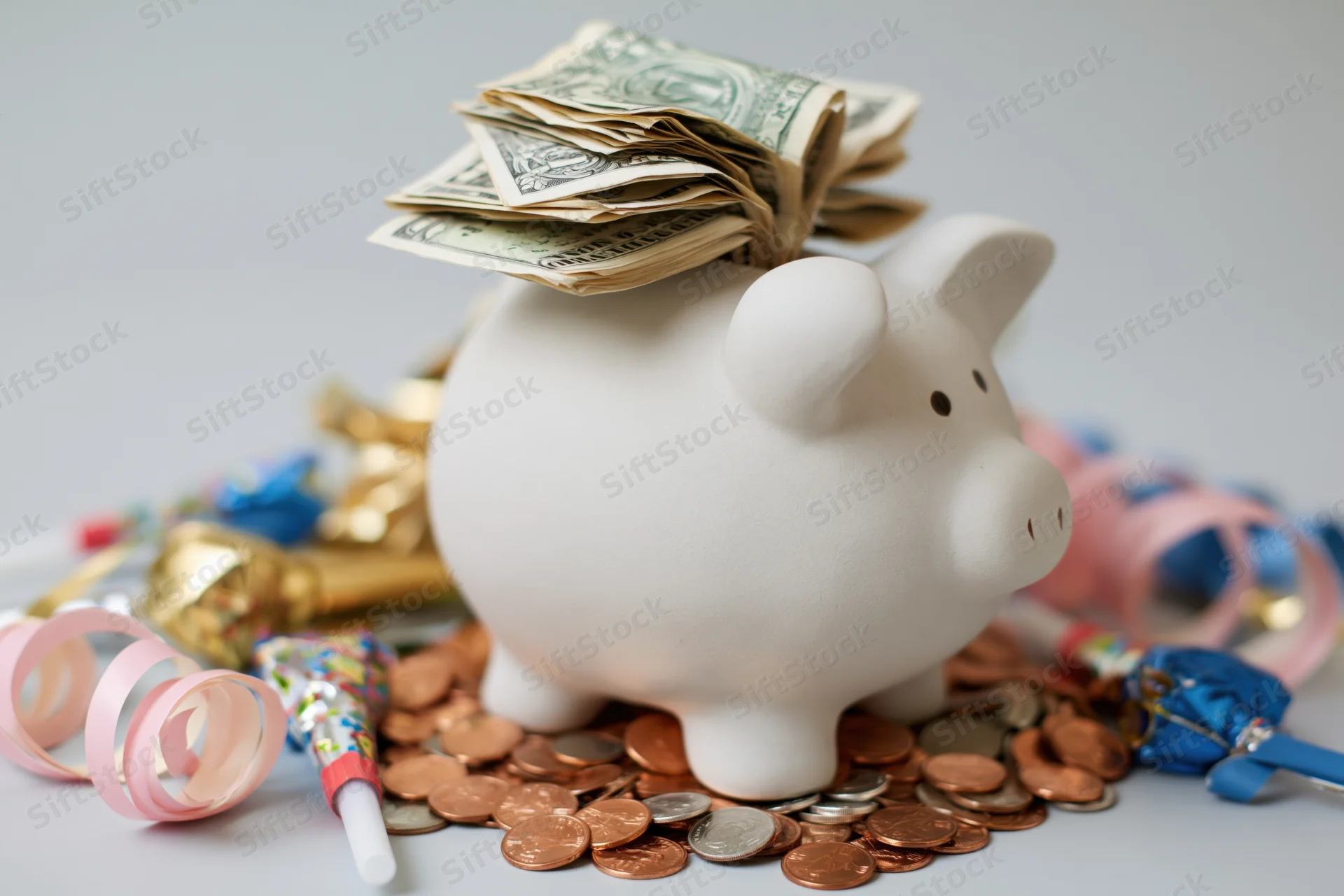 White ceramic piggy bank stuffed with folded us dollar bills surrounded by coins and party decorations on a plain surface