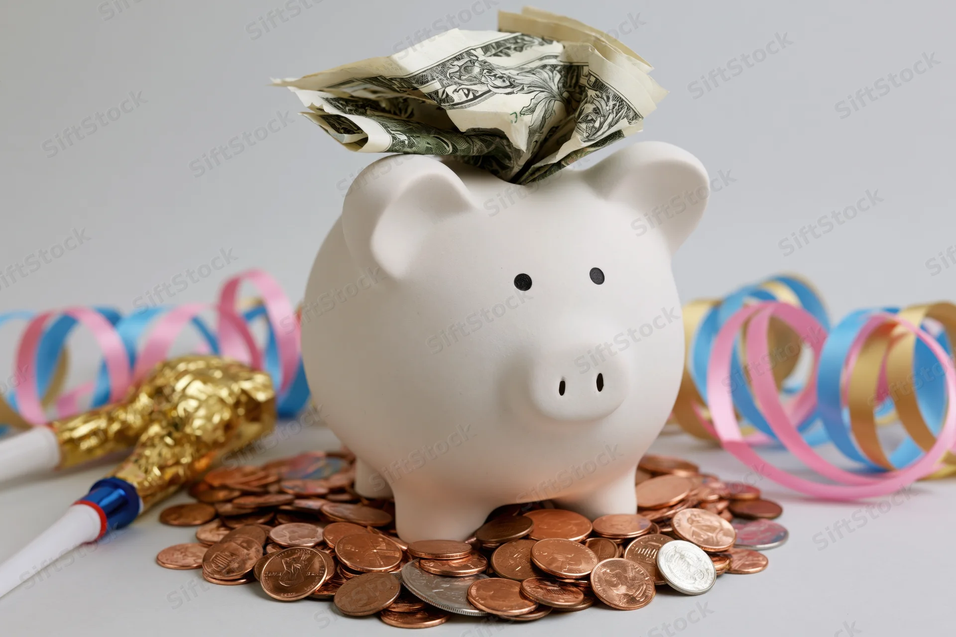 White ceramic piggy bank stuffed with folded us dollar bills surrounded by coins and party streamers on a plain surface